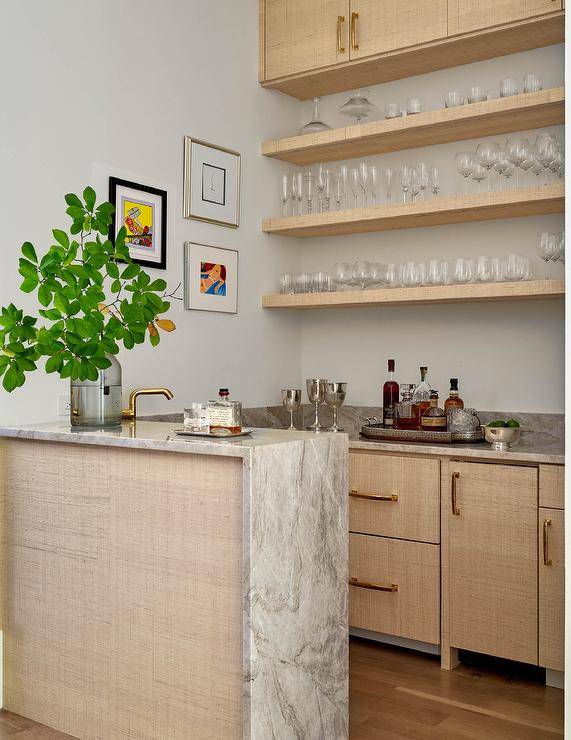 A gold raffia wet bar island is complemented with a gray marble waterfall edge countertop. Fabric wrapped cabinets adorned with brass pulls are topped with a gray marble countertop fixed beneath gold raffia wrapped floating shelves.