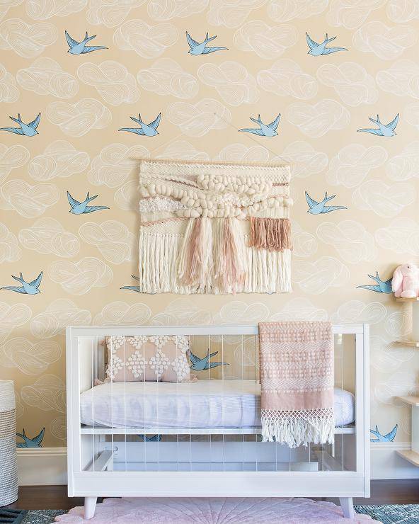 Hygge & West Daydream Wallpaper dresses up a boho baby nursery with a fun and charming finish in beige and blue. Wood and lucite nursery crib features blush pink accents on a throw fringe blanket and an accent pillow.