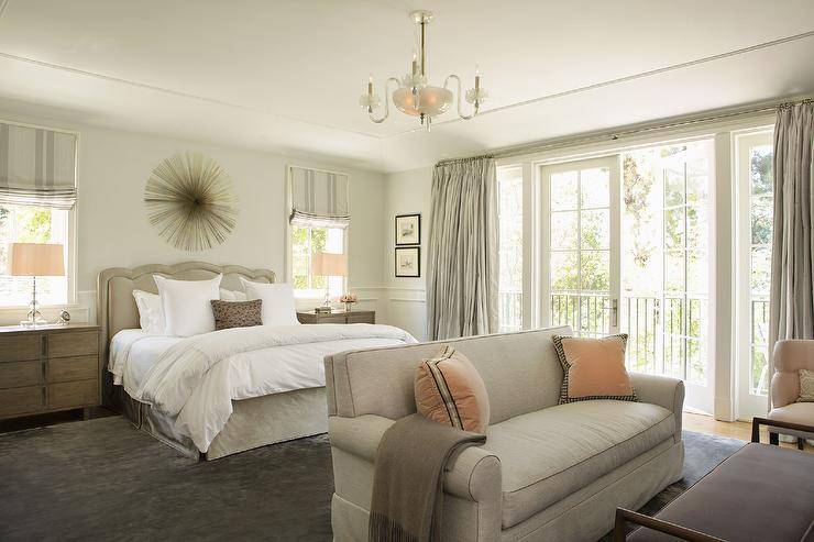 Large gray master bedroom boasts a gray bench placed on a gray rug in front of a bed boasting a gray silk pleated bedskirt and a gray silk headboard fixed under a gold sunburst decor piece. The bed is located between brown wooden nightstands topped with stacked glass lamps placed in front of windows covered in gray roman shades. A taupe love seat is complemented with orange velvet pillows and a gray throw blanket.