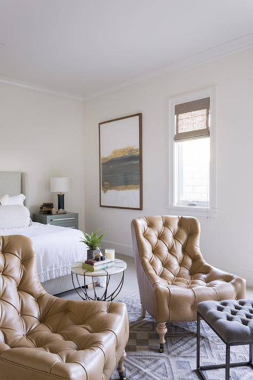 Master bedroom sitting area features caramel leather tufted accent chairs placed in front of a bed on a gray geometric rug facing a gray tufted stool.