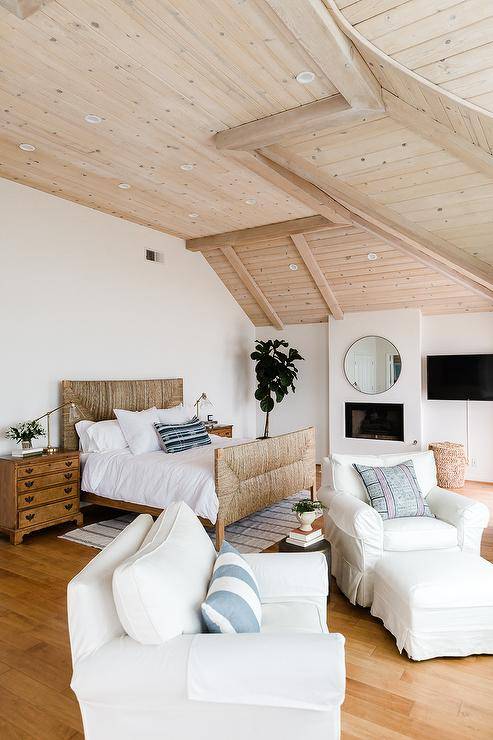 Beach cottage master bedroom boasts a light stained plank ceiling above a natural woven bed dressed with white bedding. Vintage nightstands are styled with a glass and brass task lamp adding a contemporary touch completing the rooms aesthetic.