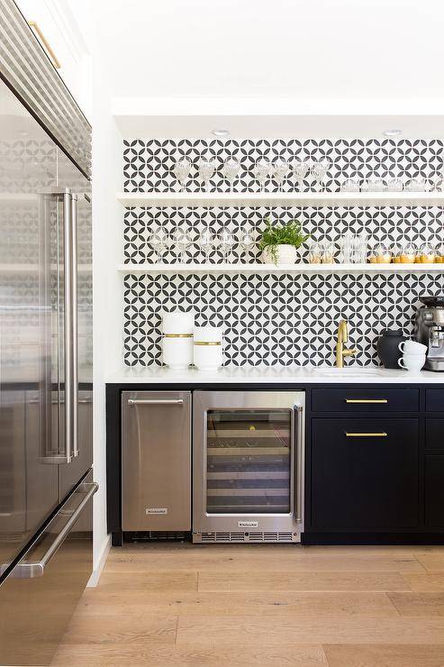 White floating shelves are fixed against black and white backsp[lash tiles in a contemporary wet bar. The shelves are fitted over black flat front cabinets adorned with brass hardware and a white quartz countertop. The countertop is mounted over a Kitchenaid wine fridge and finished with a sink and antique brass faucet.