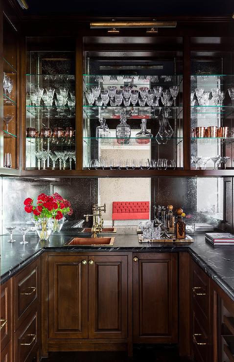 Gorgeous u-shaped wet bar boasts brown cabinets adorned with brass hardware and a beveled honed black marble countertop fixed against a mirrored backsplash. A copper hammered overmount sink matched with an antique brass wing arm faucet mounted under glass shelves.